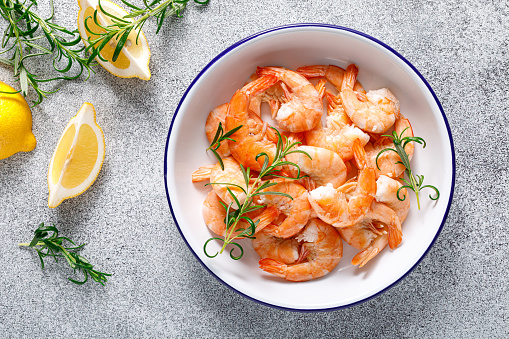 Freshly cooked shrimp served on a white plate with lemon and parsley.\nShot from above on a dark slate background. \nClose up.