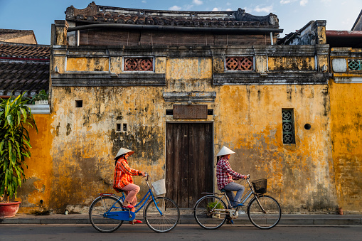 Vietnamese women ride bicycles in an old town of Hoi An city, Vietnam. Hoi An is situated on the east coast of Vietnam. Its old town is a UNESCO World Heritage Site because of its historical buildings.