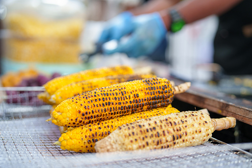 A close-up shot of grilled sweet corn on a metal tray, preparing for sale at an Asian night market.