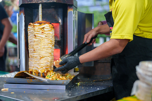 In a close-up shot, an Asian market vendor is captured preparing rotisserie by grilling chicken meat for shawarma, showcasing the sizzling and flavorful cooking process.
