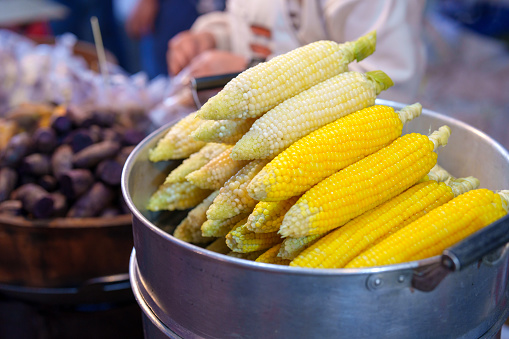 A close-up shot of steamed corn on a metal steamer tray, preparing food for sale at an Asian night market.