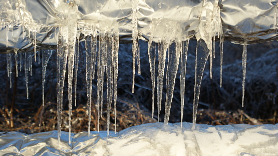 A close-up view of icicles hanging from a surface, glistening in the sunlight Below the icicles, snow is accumulated, reflecting the chilly atmosphere. In the background, dried plants partially covered by snow are illuminated by natural light, conveying a serene yet cold winter scene