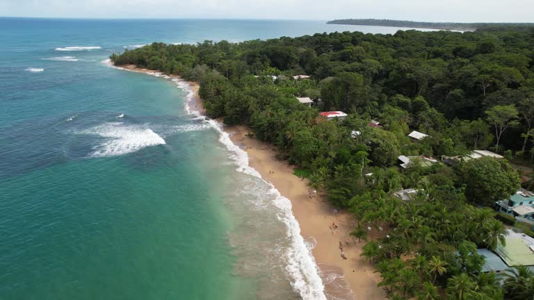 Flight over a paradisiacal beach with turquoise water bordered by a jungle with some huts.