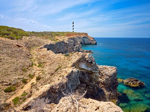 Wide-angle view of Far de sa Punta des Moscarter (also known as Faro de Portinatx), a lighthouse located on the northern coast of Ibiza, and a long stretch of the same rough coast. The dazzling warm light of a Mediterranean summer afternoon, picturesque clouds, colourful cliffs dotted with lush pine trees and Mediterranean scrub, deep blue waters as far as the eye can see. High level of detail, natural rendition, realistic feel. Developed from RAW.