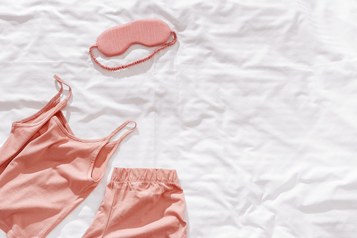 Pink woman pajamas and sleep eye mask on white bed sheet. Top view summer pyjama for sleeping. Aesthetic lifestyle flat lay photo, singlet and shorts peach colored, stylish clothes, copy space