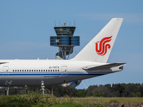 An Air China Boeing B777-39L(ER) plane, registration B-7973, has landed from Beijing as flight CA173 and taxiing to the international terminal of Sydney Kingsford-Smith Airport. She is passing the air traffic control tower.  The airport's fence and security cameras are visible.  This image was taken from Kyeemagh, Botany Bay, on a hot and sunny afternoon on 19 January 2024.