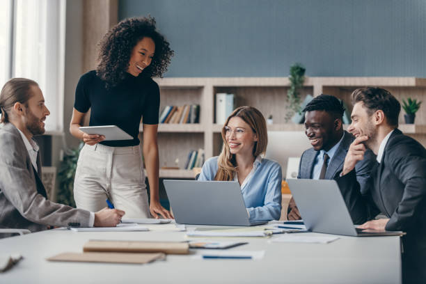 Diverse group of professionals smiling and discussing ideas during a casual office meeting with laptops and tablets.