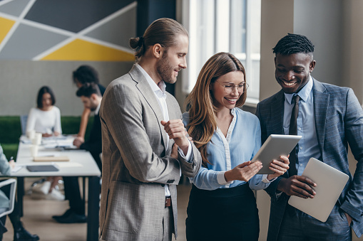 Three happy young business people looking at digital tablet while their colleagues working on background