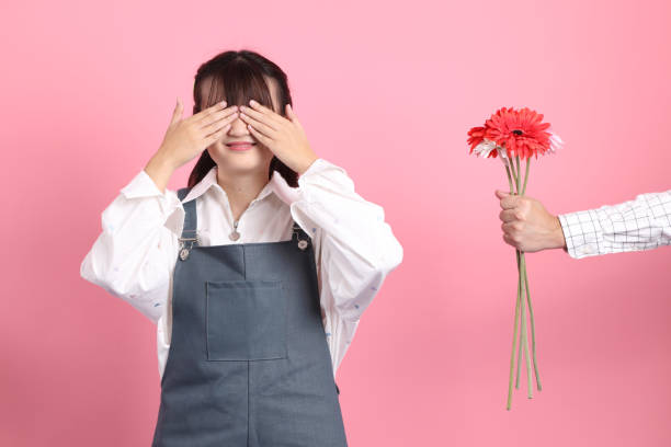 Valentine's Day Man presenting flowers to cheerful lovely young asian woman in overalls casual clothes with gesture of receiving isolated on pink background. St Valentine's Day, Women's Day, Birthday unusual couple stock pictures, royalty-free photos & images