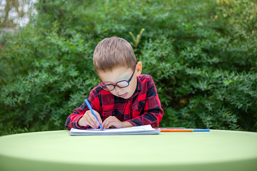 Kid draws in the garden (outdoors). He wears glasses and an eye patch (occluder) to prevent amblyopia and strabismus (squint, lazy eye). The preschooler and school students vision diseases problem.