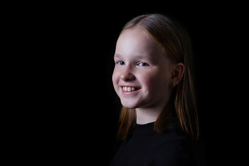 Portrait of beautiful white girl with freckles on dark black background. Close up portrait of young happy smiling girl in low key.