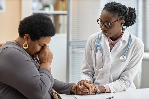 Medium shot of African American woman doctor in glasses supporting tearful female patient while holding hand and looking at her