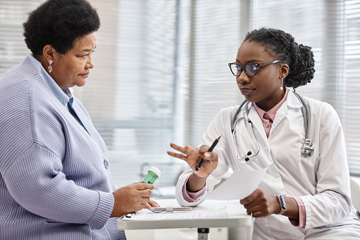 Senior African American female patient holding drug bottle while listening to doctors instructions about medication administration in clinic office