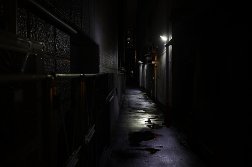 A silhouetted person walks through the shadows of and alleyway towards camera.