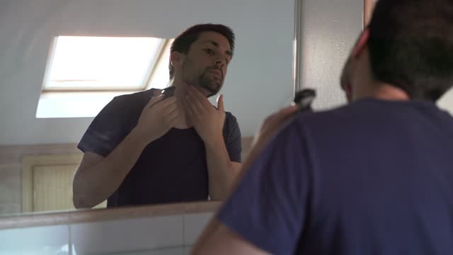A man stands in front of a mirror as he razor his beard