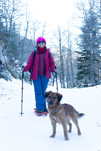 portrait photograph of snowshoeing woman and her adorable dog looking at the camera in the woods in the wintertime