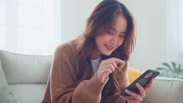 Happy young asian woman surfing social media on mobile phone, enjoying amazing news , Female having fun playing games chatting in social media sit on couch at home