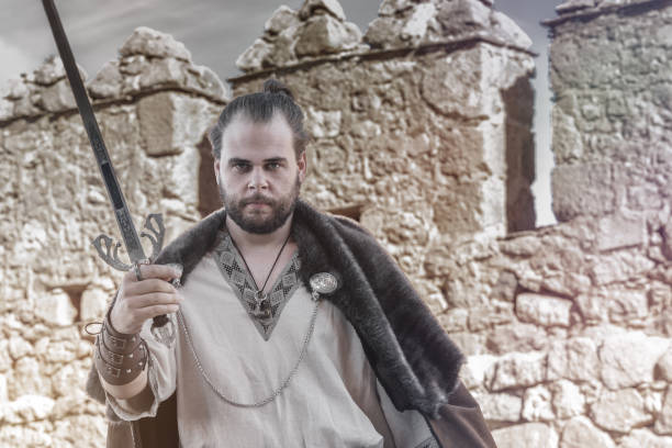 A medieval warrior holds a sword, standing firm in front of a stone fortress ruin under a cloudy sky. exterior of the Calahorra castle