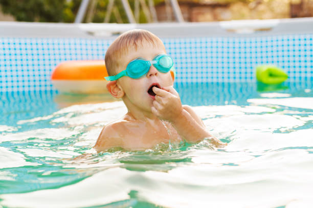 Cheerful young boy with swim goggles emerges from the water in a pool, mouth open in mid-talk or laughter, enjoying a refreshing swim.