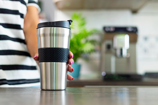 Close-up of a hand holding a stainless steel travel mug, ready to enjoy a hot drink from the comfort of a well-lit home kitchen.