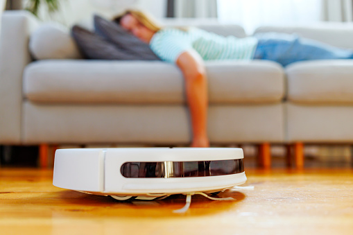 A peaceful scene of a woman taking a nap on a sofa while an autonomous robotic vacuum cleaner tidies the living room, symbolizing modern home convenience.
