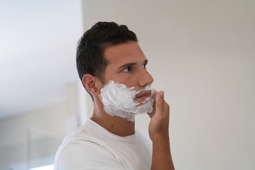 A young man is applying some shaving foam on his face at the  bathroom of his home
