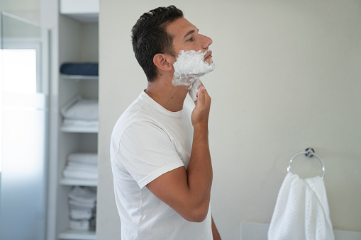 A young man is applying some shaving foam on his face at the  bathroom of his home
