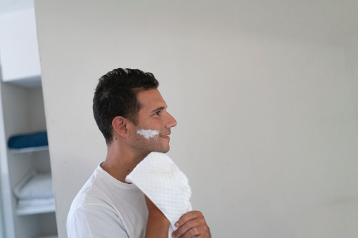 A young man is drying his face after shaving with a towel at the  bathroom of his home