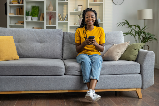Shot of a young woman using a smartphone and wireless earbuds on the sofa at home