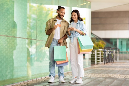 Cheerful millennial european couple with bags enjoy shopping app on smartphone, credit card in mall, full length. Sale for shopaholics, discount, buy order, cashback and money finance