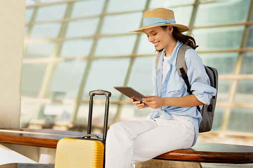 Cheerful young european woman in hat with suitcase and backpack, typing on tablet, enjoy journey on station, airport. App for tourist, business trip, active lifestyle, work and vacation