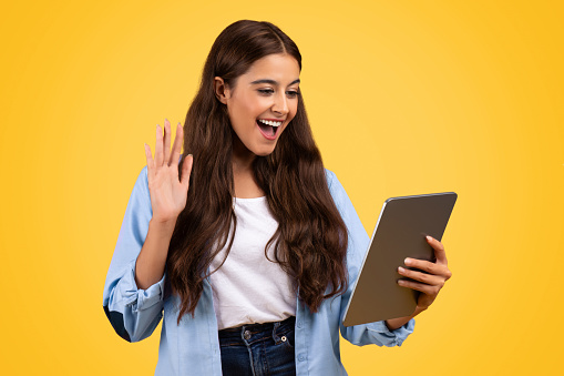 Smiling pretty european woman student, use tablet, waving hand, isolated on yellow studio background. Education, online lesson, education project remote, video call with teacher