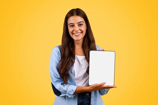 Positive european young student lady show tablet with empty screen, isolated on yellow background, studio. Study remotely recommendation, education and online lesson, attention courses app