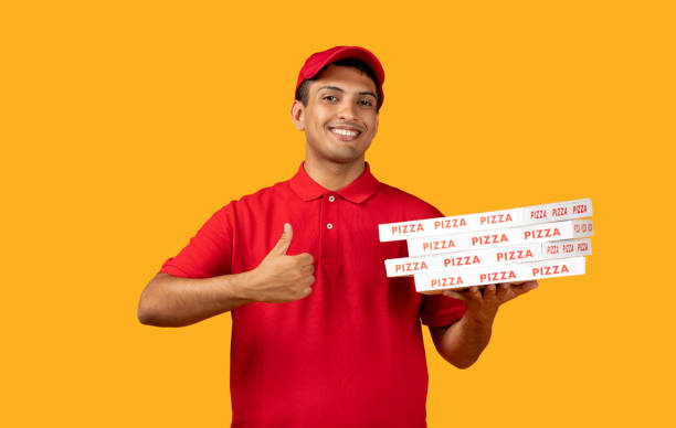 Pizzeria Delivery Offer. Mexican Courier Guy Holds Stack Of Pizza Boxes Gesturing Thumbs Up On Yellow Studio Background. Deliveryman Advertising And Approving Food Delivering With Like