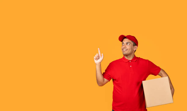 istock deliveryman holding cardboard box pointing finger towards copy space, studio 1948033852
