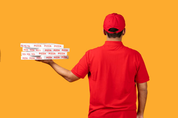 Rear view of courier man holding stack of takeaway pizza boxes from restaurant over yellow studio backdrop. Meal ordering and delivery concept. Shot of unrecognizable pizzeria deliveryman