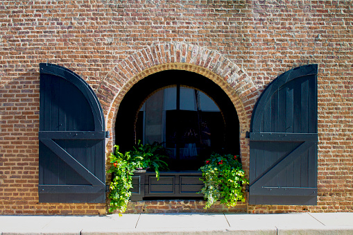 An arched window with black wooden shutters and a black flower box against a flat brick wall in Charleston, SC