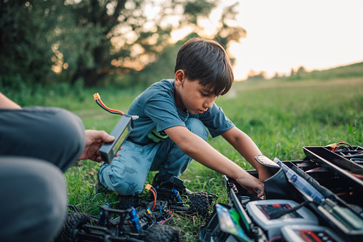 Picture of a boy kneeling over a chassis of his toy monster truck and taking out the tool out of the toolbox. There is the father's hand holding the battery box with wires, handing it to son.