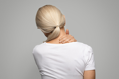 Sick mature blond woman suffering acute neck pain and strain, unrecognizable older lady rubbing sore area with hand, massaging neck while standing over grey studio background, copy space