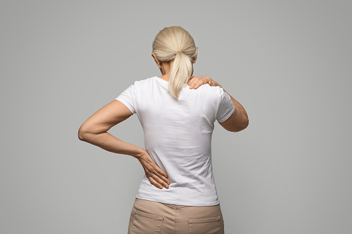 Unrecognizable mature blonde woman suffering neck and back pain, rear view of sick tired lady rubbing her painful body, suffering from office syndrome, standing on grey studio background, copy space