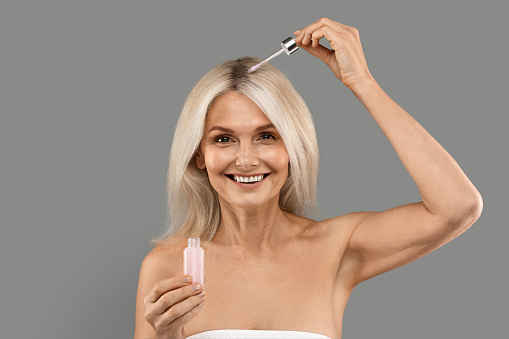 Beautiful Mature Woman Applying Hair Serum With Droplet, Attractive Senior Lady Wrapped In Towel Moisturising Sculp Skin And Looking At Camera, Standing Over Grey Background In Studio, Copy Space