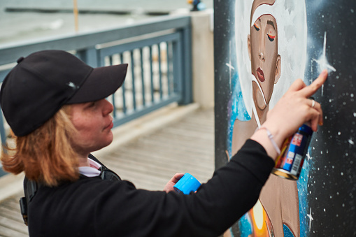 Female artist in black cap is painting picture with paint spray can spraying it onto canvas at outdoor street exhibition, side view of female art maker