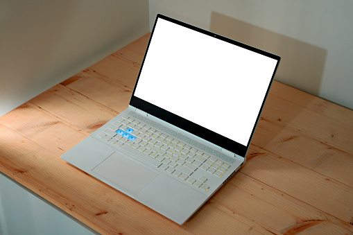 A template of a white laptop on a desk. a slim gaming notebook with white screen on desktop. display is changeable and easy to use mockup on a wooden table and white wall background. RGB keyboard and minimal design environment.