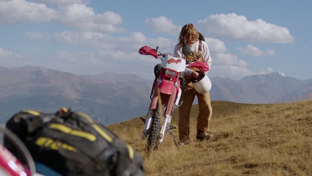 A male motorcyclist removes his helmet near a vintage off-road enduro motorcycle against a backdrop of mountains and sky with clouds. Riding a motorcycle in the mountains. A man in a suit in the mountains of Appalachia. Redneck in natural environment.