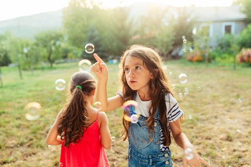 Two cheerful girls having fun catching soap bubbles in the yard