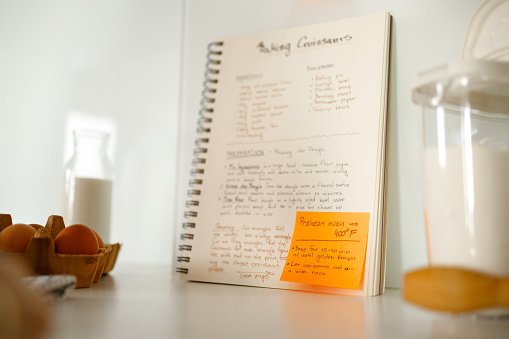 The photo captures an open recipe book with detailed instructions for making croissants, accompanied by a sticky note with additional baking tips, set in a kitchen scene with blurred ingredients in the foreground.