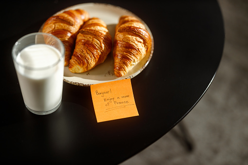 A plate of freshly baked croissants paired with a glass of milk on a black table, complemented by a sticky note that reads \