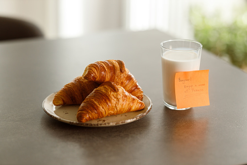 A plate of freshly baked croissants paired with a glass of milk on a black table, complemented by a sticky note that reads \