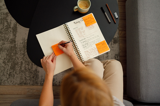 A woman is engrossed in writing a recipe in her notebook, seated comfortably on a couch. She uses sticky notes for additional cooking instructions, with a cozy home environment that includes a cup of coffee on the coffee table.