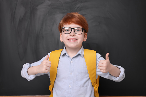 Smiling schoolboy in glasses showing thumbs up near blackboard